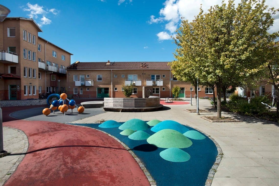Ett torg med färgglad lekplats i förgrunden. I bakgrunden finns flerfamiljshus i tegel, ett på fyra våningar samt ett på två våningar