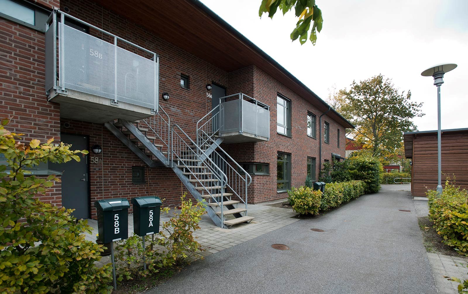 En tegelbyggnad med en utomhustrappa som leder upp till en ingång. Det finns gröna buskar runt byggnaden och brevlådor vid foten av trappan. 