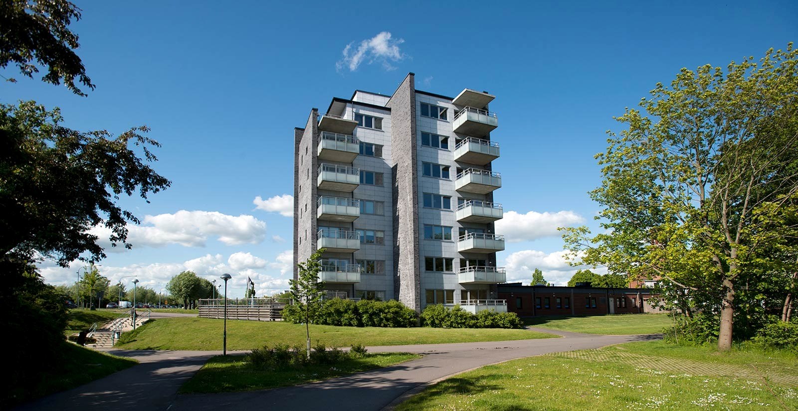 en modern flervåningsbyggnad omgiven av grönska under en klar himmel. Byggnaden har en grå fasad, s