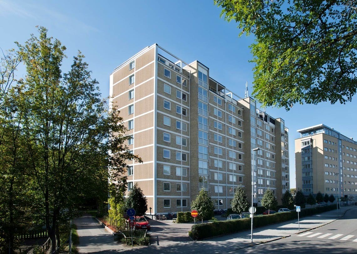 En stor bostadsbyggnad omgiven av grönska och en klarblå himmel. Byggnaden är ljusbrun med många fö