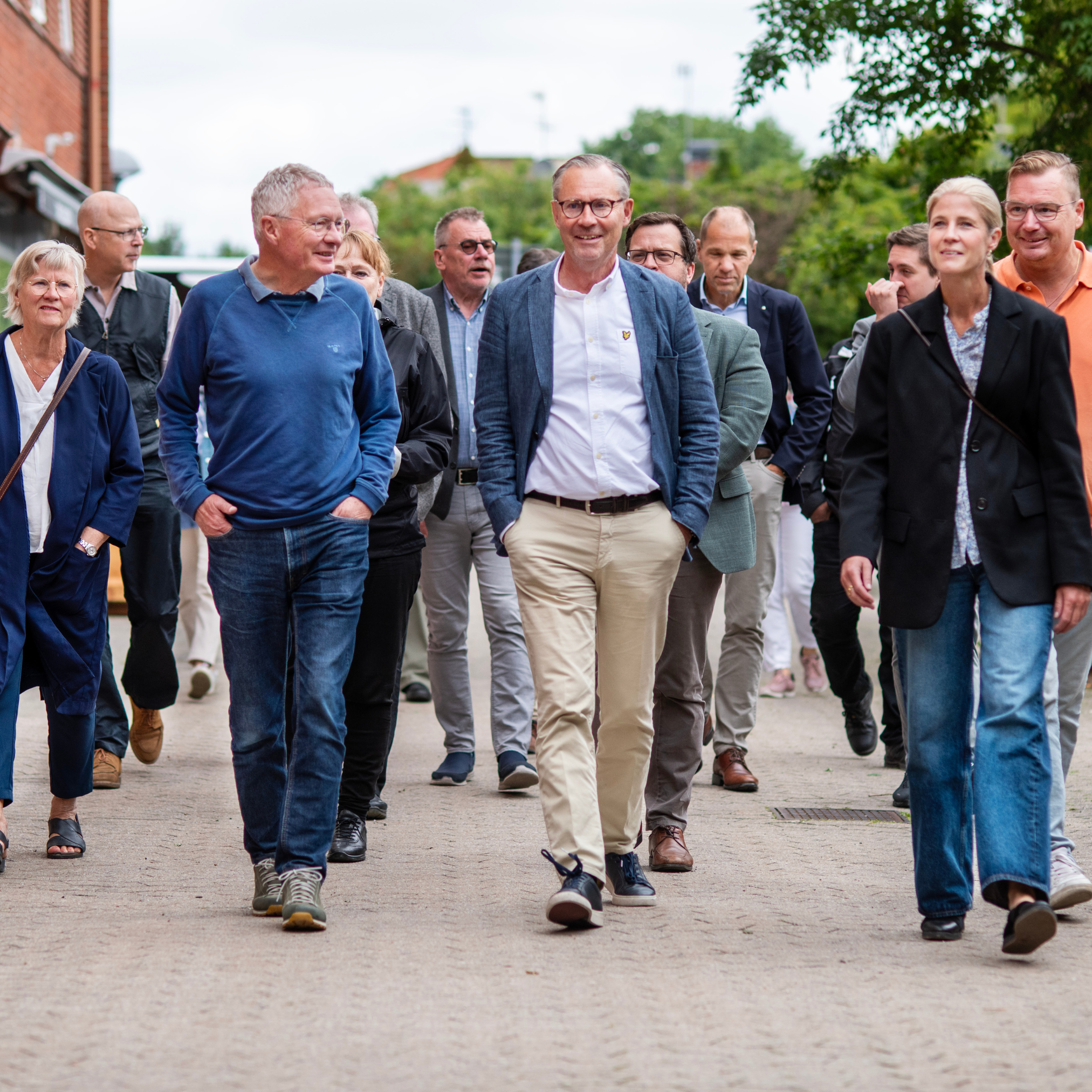 Leende personer promenerar och pratar med varandra. Hus skymtas i bakgrunden