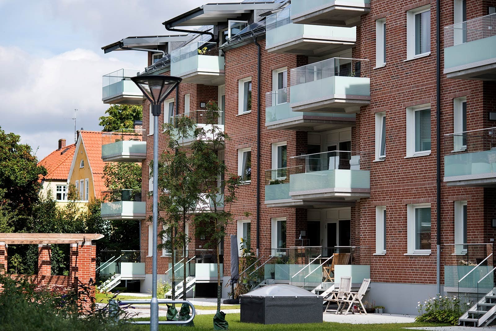 En modern bostadsbyggnad med röda tegelstenar och flera balkonger med glasräcken. En välskött trädg