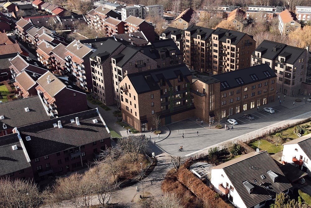 En flygbild av ett urbant område med en variation av byggnader. Det finns flera flervåningshus för bostäder med mörkbruna och röda fasader och sadeltak. 