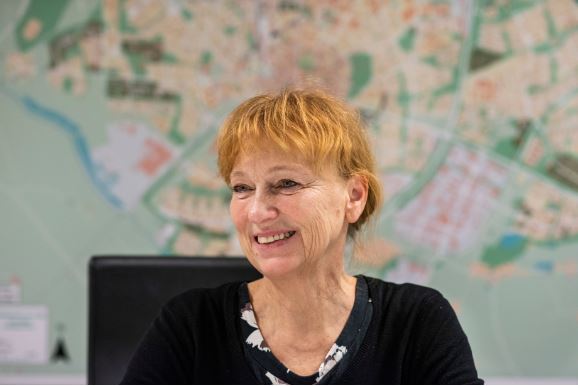 Leende kvinna med rött uppsatt hår och lugg. Vägg med karta skymtar i bakgrunden.