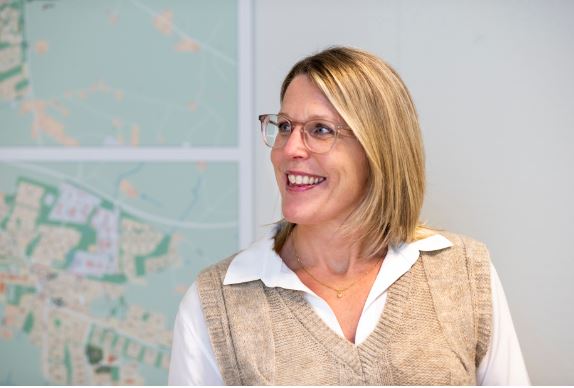 Leende kvinna led ljus lång page och glasögon. Vägg med karta skymtar i bakgrunden. 
