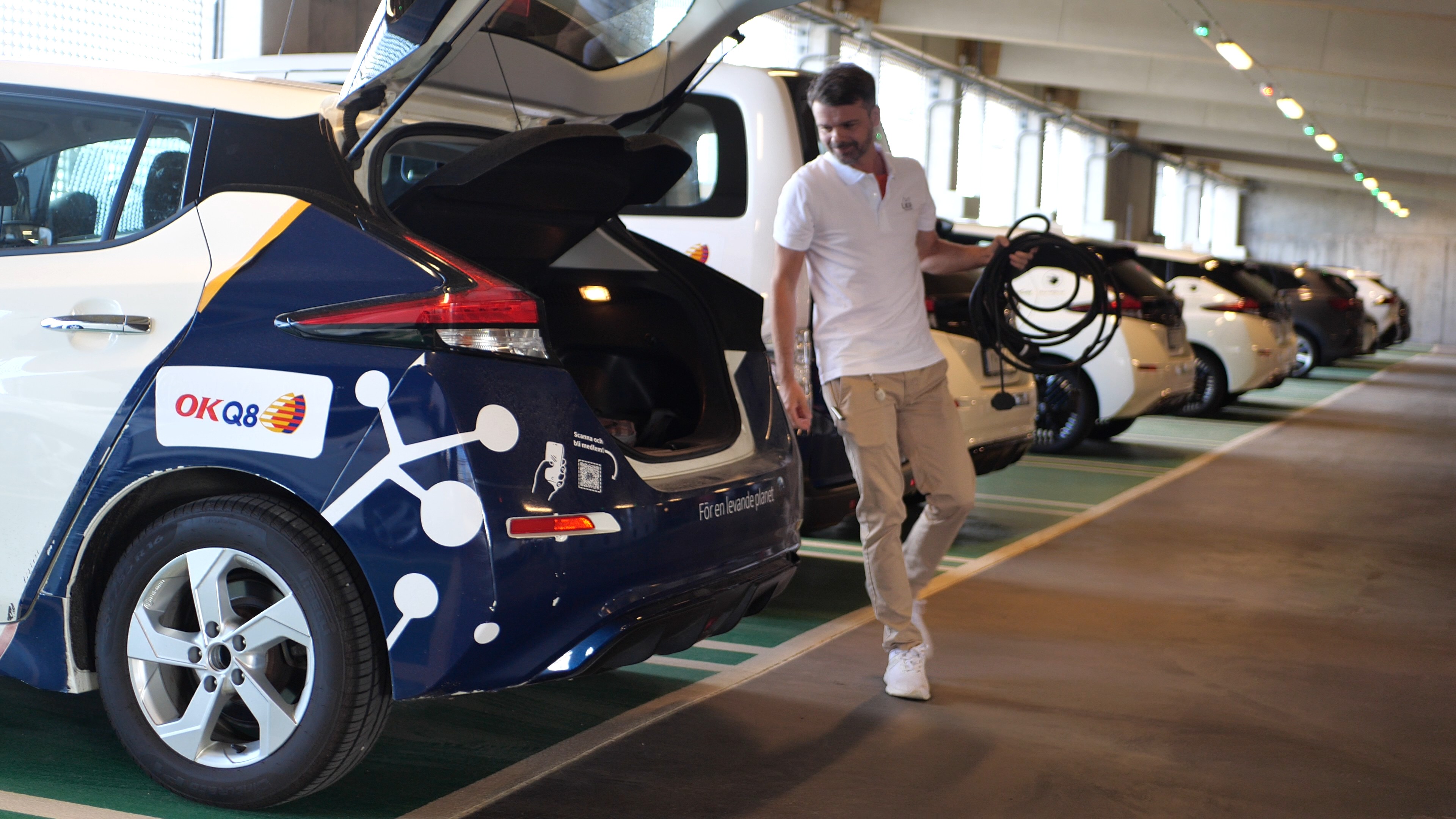 En person packar ner en laddkabel i elbilens bageutrymme. OKQ8-loggan syns på bilen.