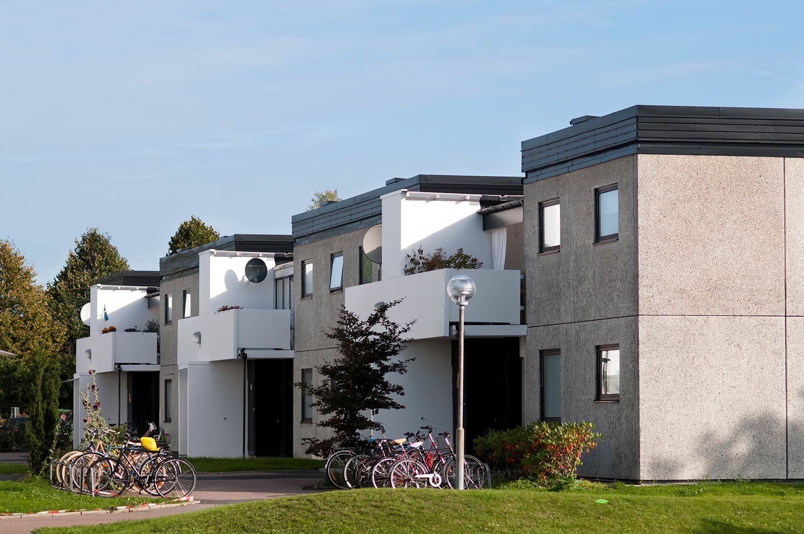 En rad hus med minimalistisk arkitektur i vitt och grått, omgivna av en välskött gräsmatta. Flera cyklar är parkerade framför husen och en gatlykta står på gräsmattan. 
