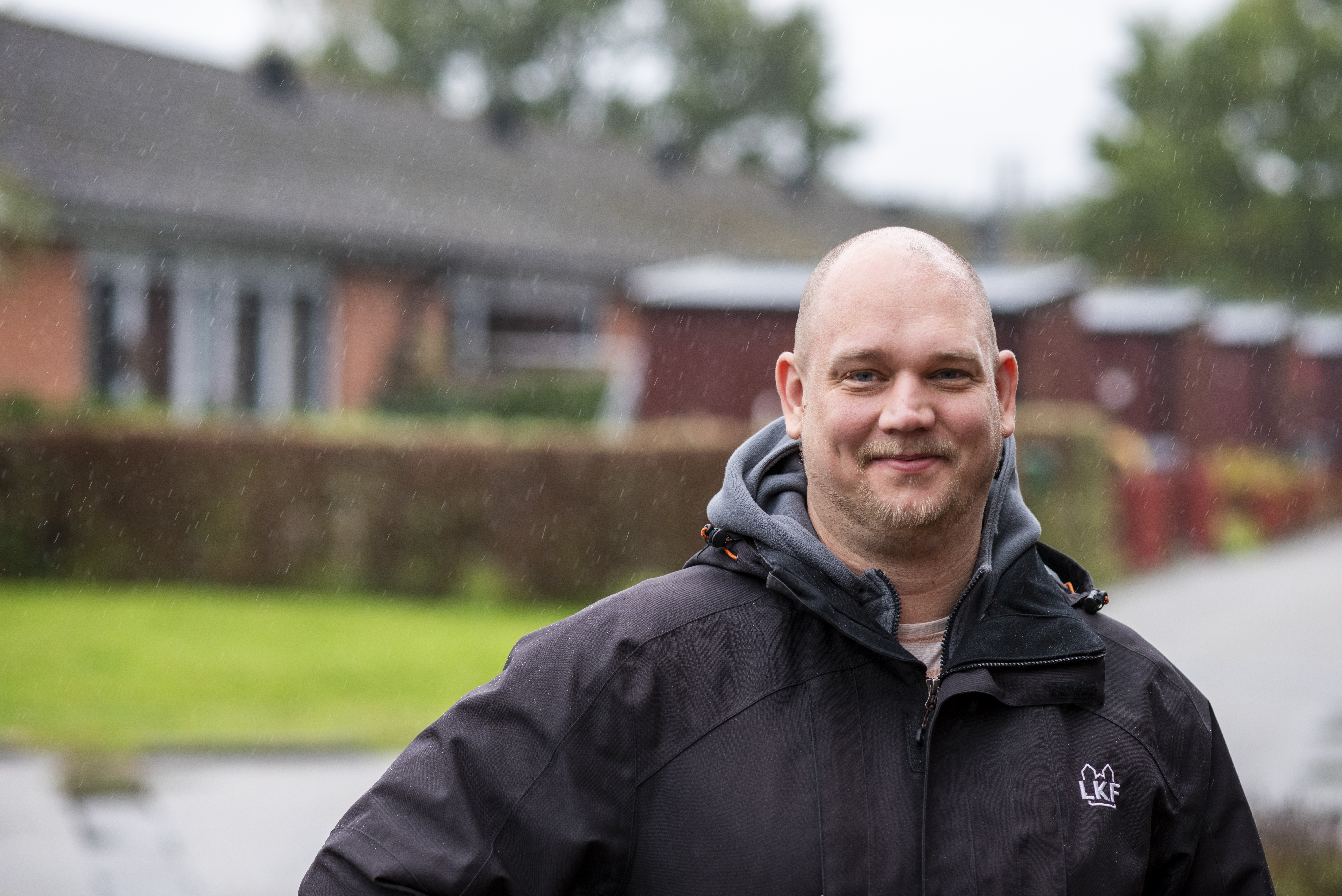 Leende man med svart jacka med "LKF" text i vitt. Radhus och gräsmatta skymtar i bakgrunden.