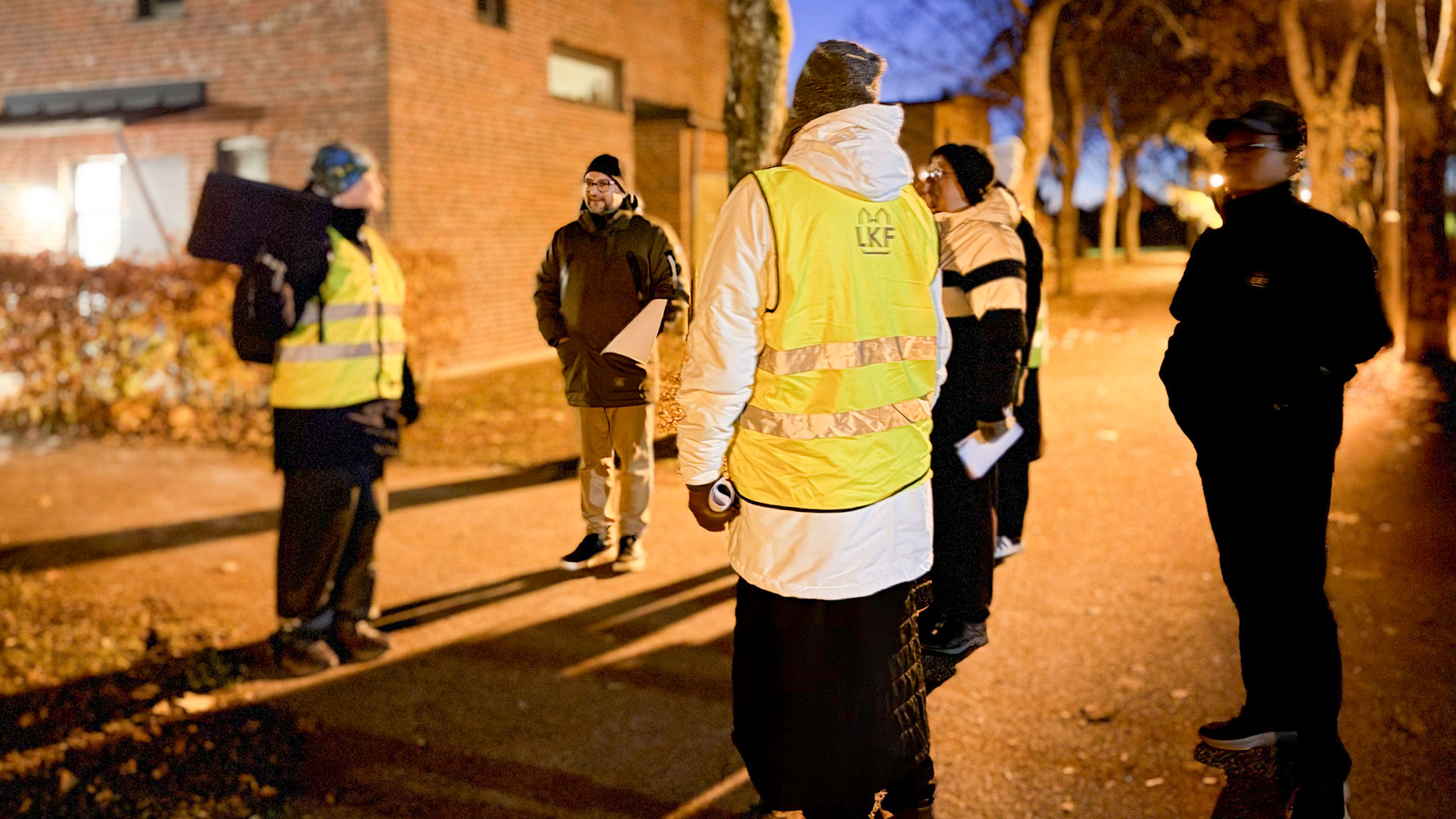 Fyra personer, två med reflexvästar står på innergård i Nöbbelöv i kvällsmörkret där gatljuset lyse