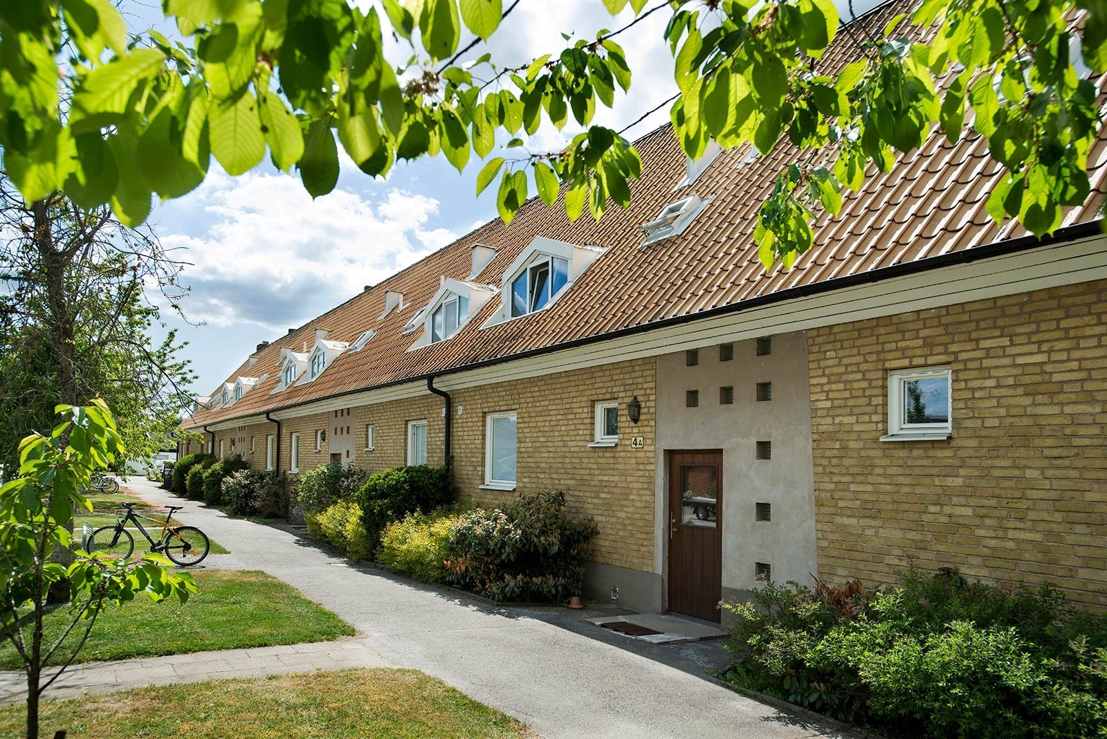 En rad sammanlänkade hus med gula tegelväggar och bruna tak. Varje hus har små fönster och en trädörr vid entrén. En stig leder fram till varje hus, och en cykel är parkerad på stigen. Husen är omgivna av välskött grönska, inklusive buskar och träd. 