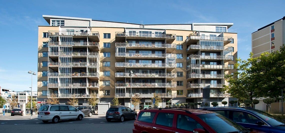 En modern flervåningsbyggnad med balkonger, omgiven av parkerade bilar under en klar himmel. Fasaden är beige, och balkongräckena är metalliska med horisontella linjer. 