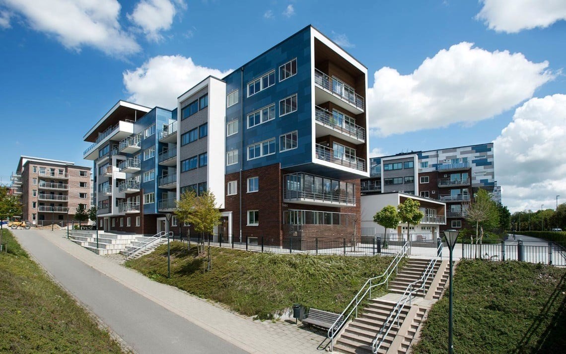 En modern bostadsbyggnad med flera våningar och en blandning av blå, vit och tegelröd fasad. Varje lägenhet har en balkong, och det finns en asfalterad gångväg som leder till byggnaden. Området är omgivet av en välskött gräsmatta, små träd och buskar. 