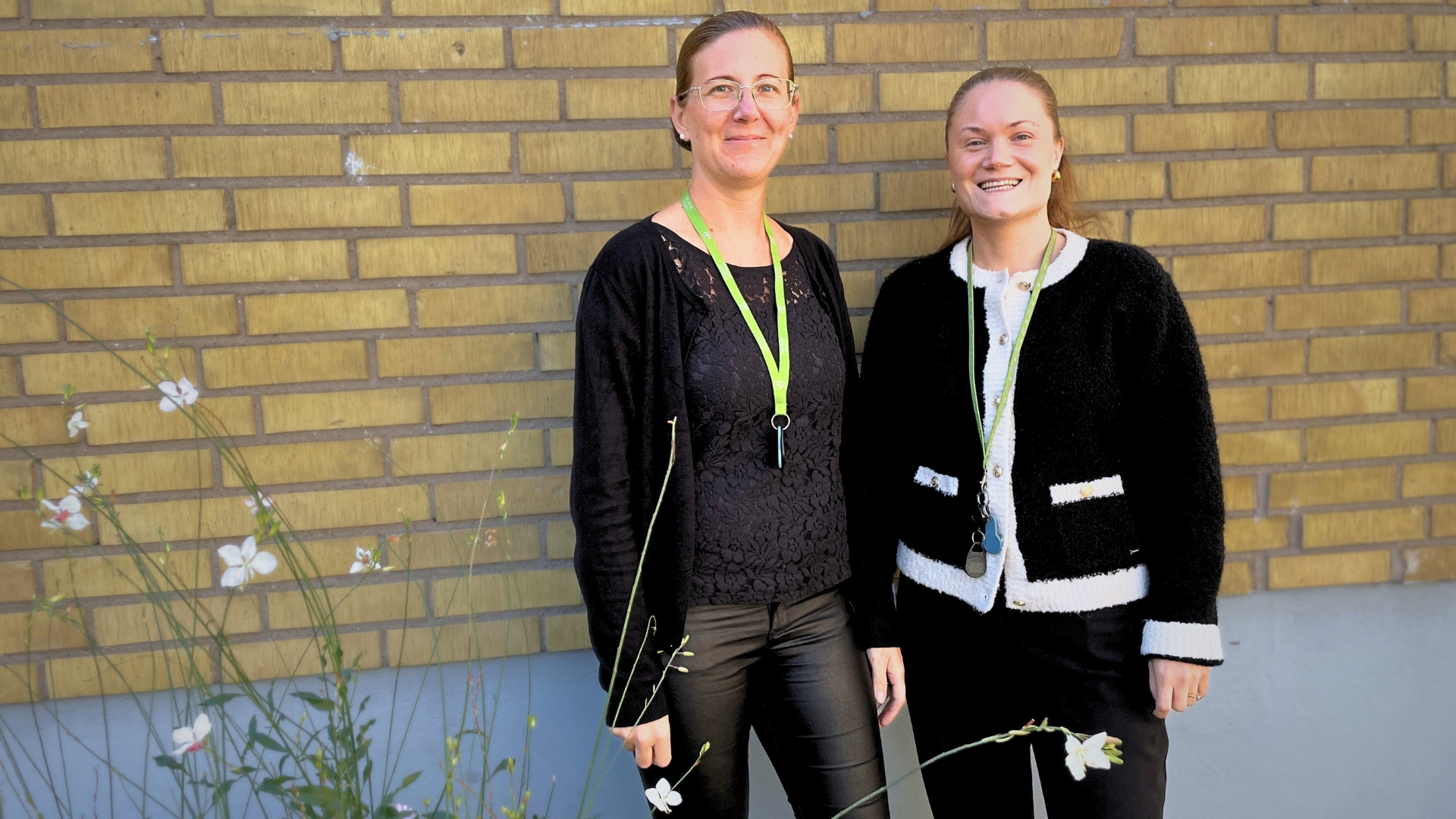 Två leende kvinnor i svarta kläder och gröna LKF-nyckelband som står mot en guld tegelvägg. Blommor 
