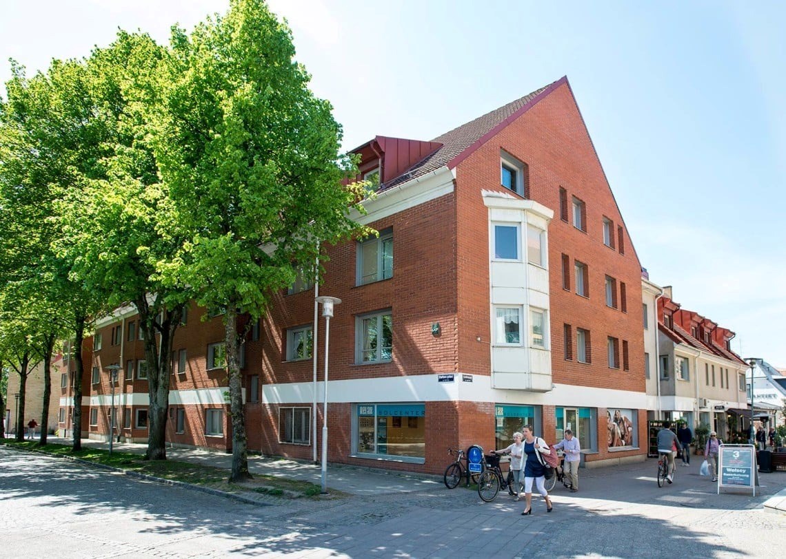 En modern tegelbyggnad med flera fönster och en ingång. Framför finns en stadsgata på en solig dag med klar himmel. En stor, frodig grön träd finns till vänster i bilden. Människor går längs trottoaren.