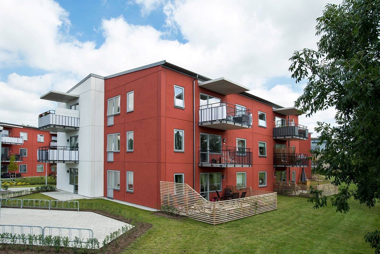 En modern bostadsbyggnad med tre våningar, målad i en röd färg med vita accenter runt fönstren och balkongerna. Varje lägenhet har en liten balkong med svarta räcken. Byggnaden är omgiven av en välskött grön gräsmatta och några träd som tillför naturlig skönhet till området