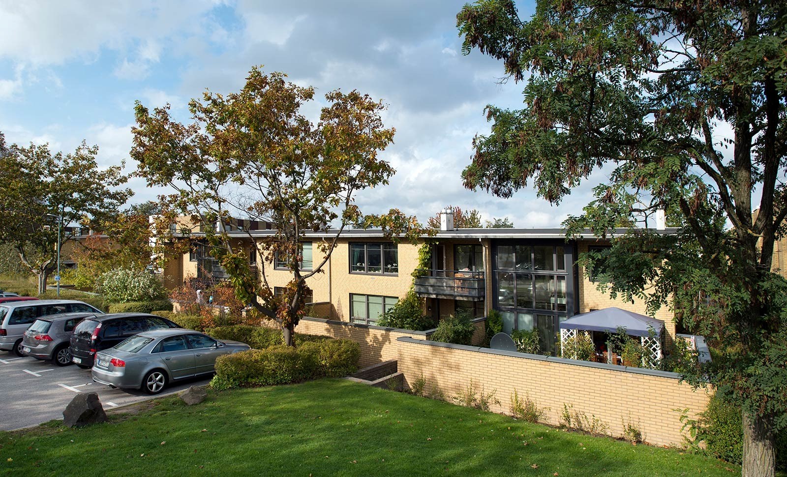 En modern byggnad omgiven av grönska och parkerade bilar. Byggnaden har stora fönster och en balkong. På en av uteplatserna finns ett parasoll. Framför byggnaden syns en välskött gräsmatta och buskage.