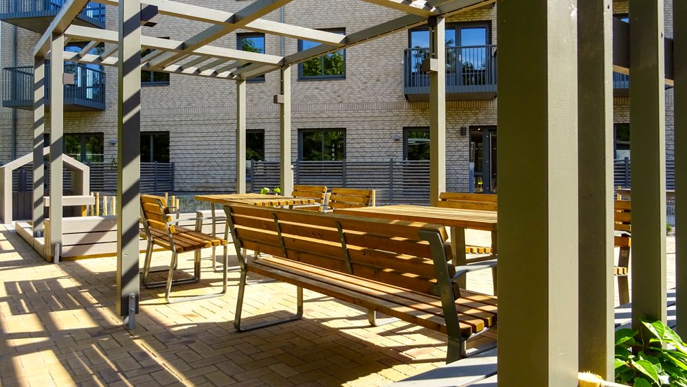 Sittplatser under en pergola. I bakgrunden skymtar den lilla lekplatsen med lekstuga och sandlåda. Från innergården