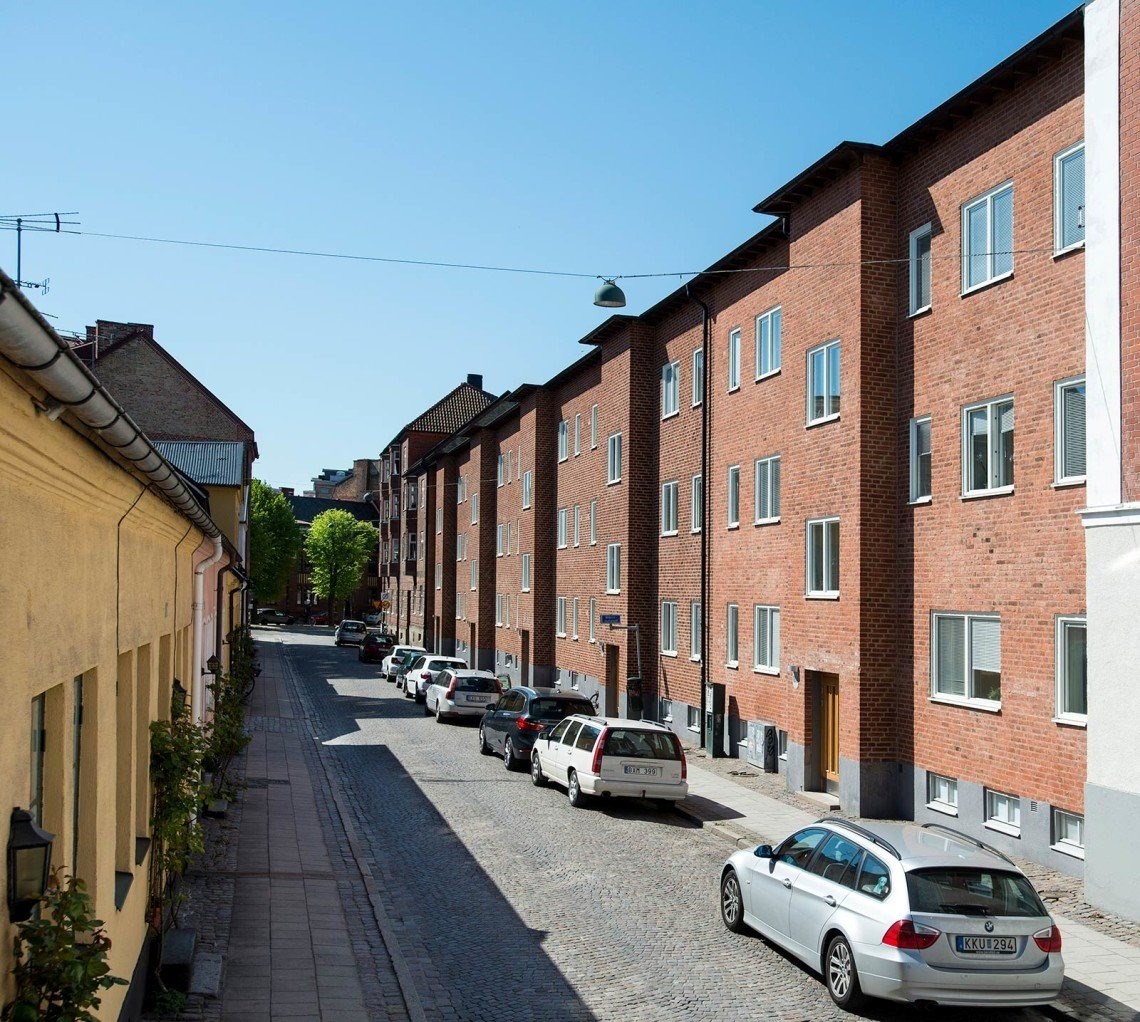 En lugn och solig stadsgata. På ena sidan syns röda tegelbyggnader med jämnt fördelade fönster. Till vänster finns en byggnad i annan färg, möjligen äldre än de röda tegelbyggnaderna.