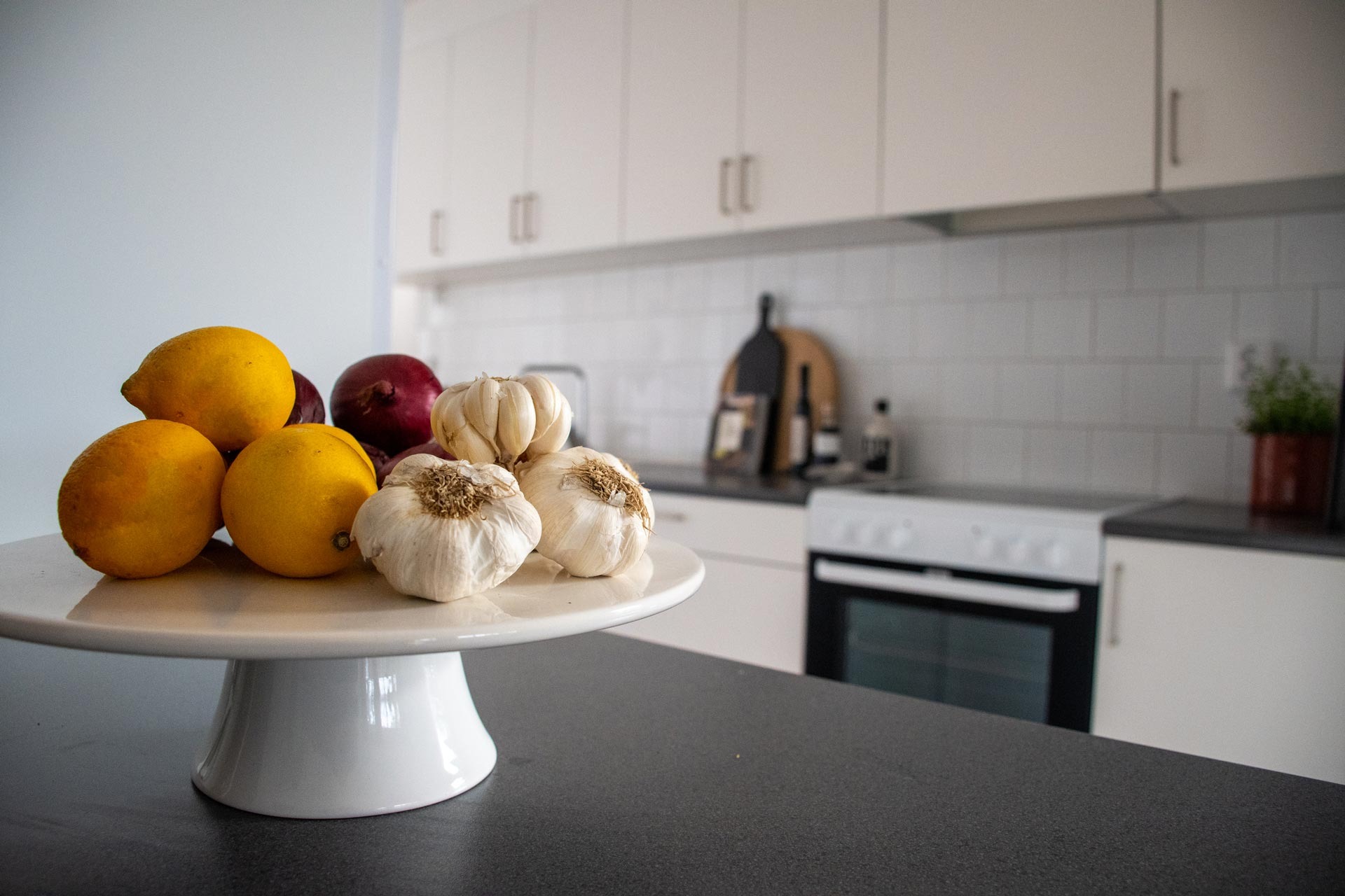 En vit keramisk kakfat på en mörk köksbänk. Ovanpå stället finns en samling gula citroner och vita vitlöksklyftor, med ett ensamt rött äpple synligt bakom dem. I bakgrunden finns ett modernt kök med vita skåp och en inbyggd ugn.