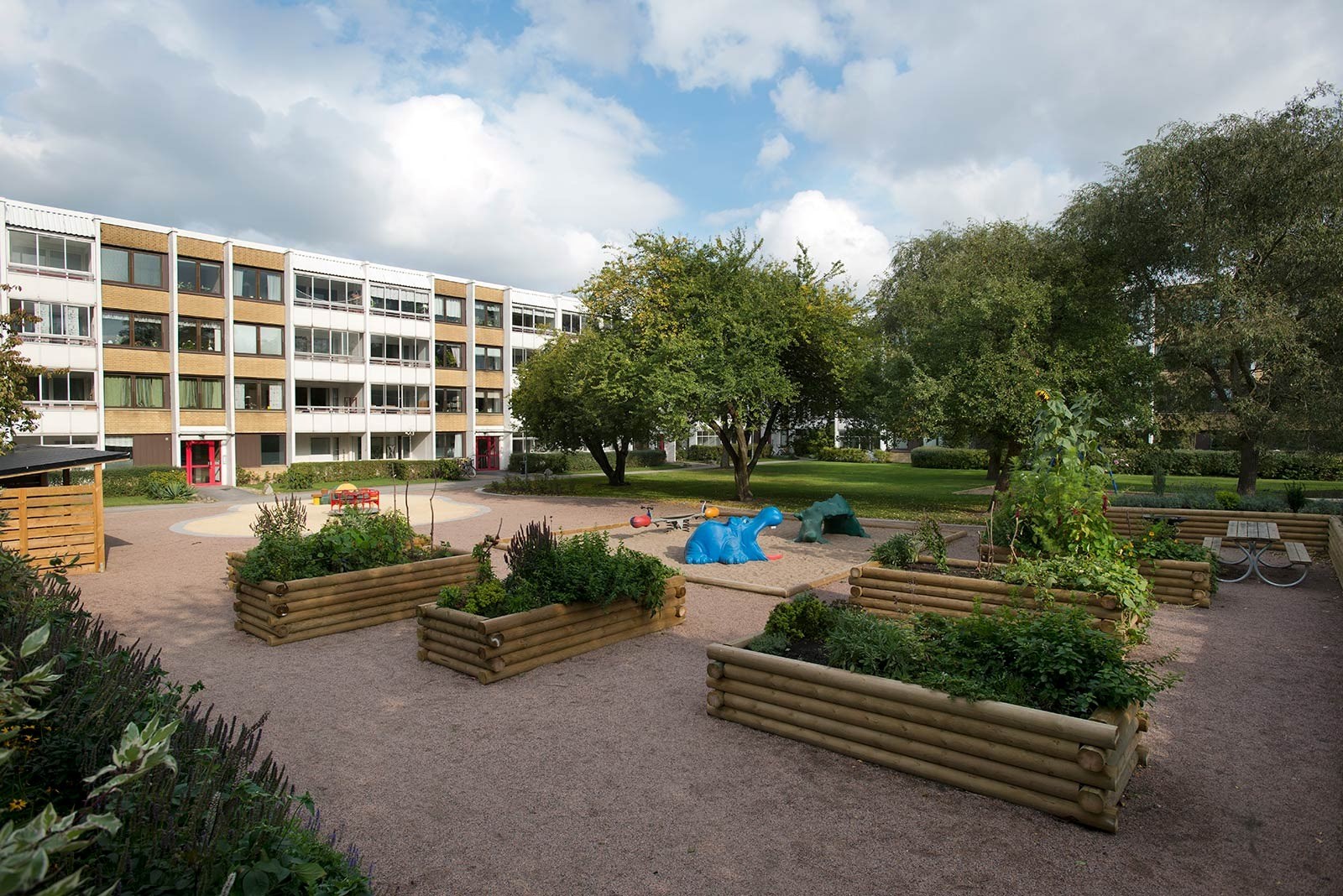 En flervåningsbyggnad med flera fönster och balkonger. Byggnaden har en röd entrédörr och är omgiven av en välskött innergård. Innergården har träplanteringar med gröna växter och buskar. En lekplats med färgglada lekredskap är placerad på en grusbelagd yta. Dessutom finns det bänkar runt om.