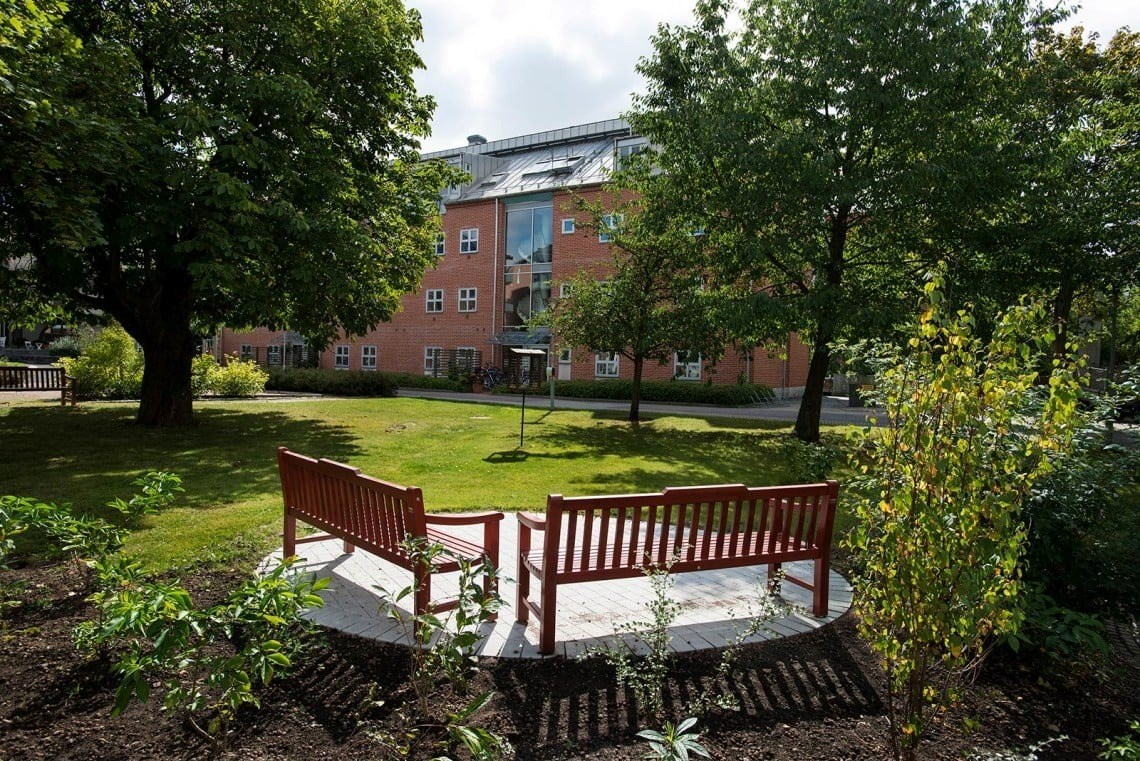 En utomhusmiljö med en röd träbänk placerad på en vit plattform, omgiven av grönska. I bakgrunden syns ett stort flervåningshus med tegelfasad.