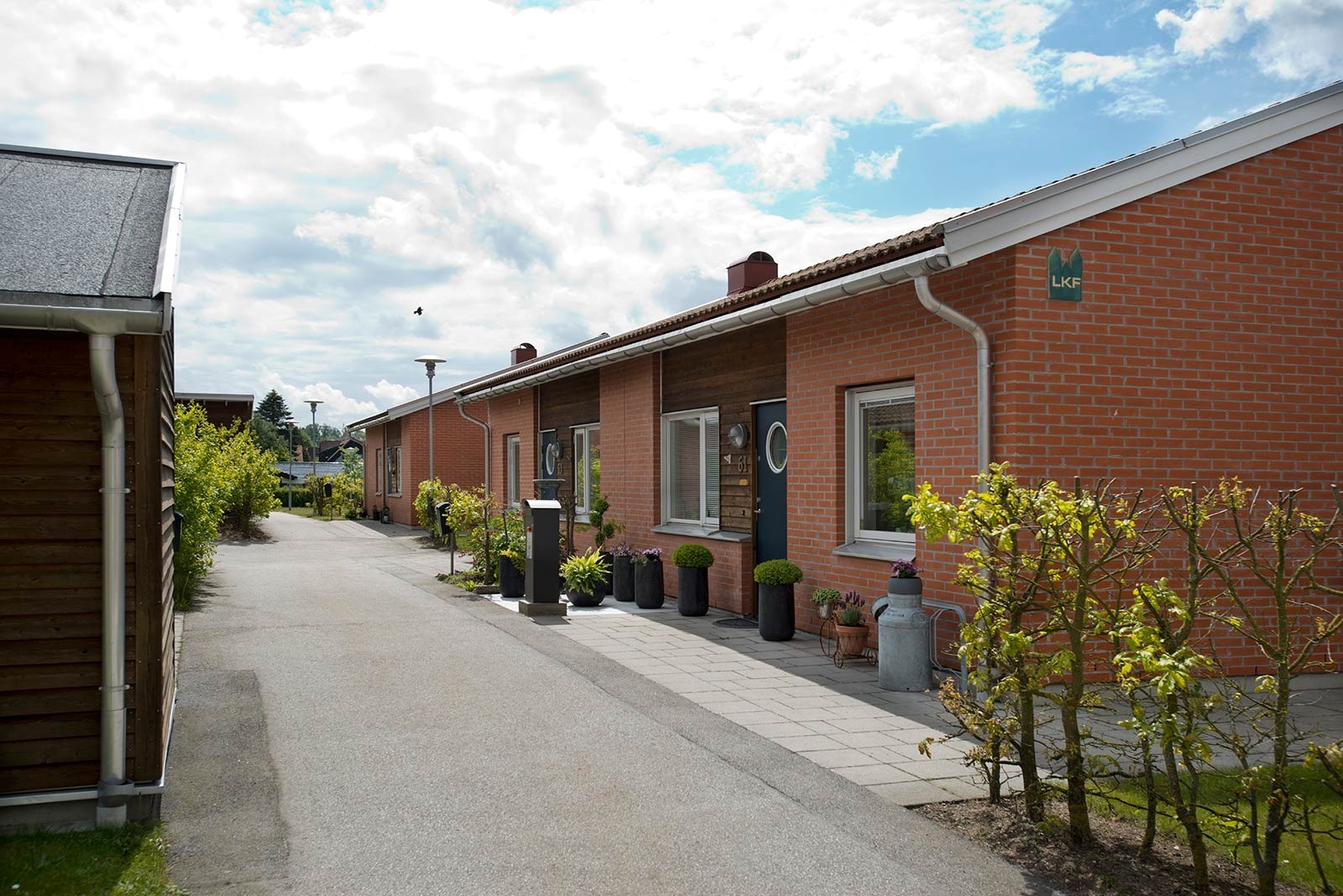 En rad tegelröda enfamiljshus med vita fönster och dörrar. Varje hus har en liten trädgård framför med krukväxter och små träd. En betonggång leder fram till varje husentré.