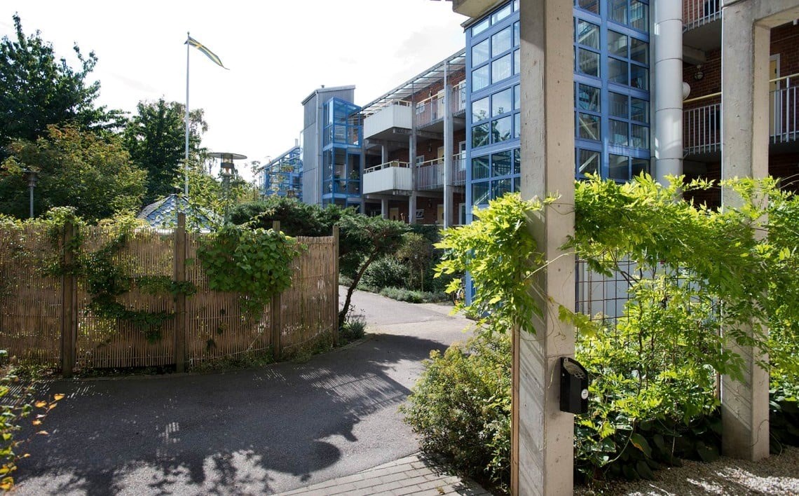 En lugn och pittoresk utomhusmiljö med moderna bostadshus i bakgrunden. En grusväg leder fram till en grind omgiven av gröna växter och träd. Bostadshusen har flera balkonger, stora fönster och är konstruerade med en kombination av tegel och blå material som ger dem en modern look.