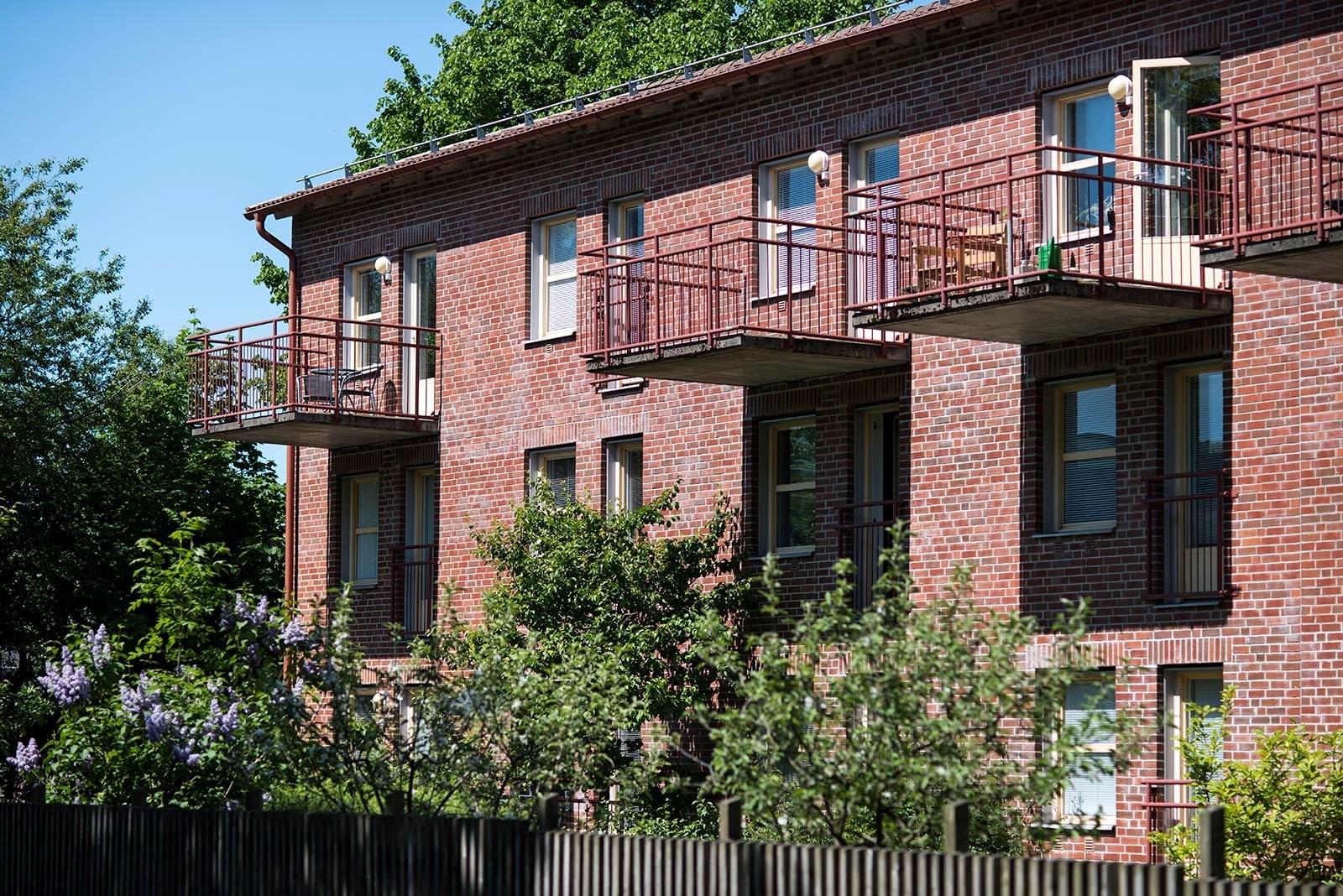 En tegelbyggnad med balkonger, omgiven av grönska under en klar himmel. Byggnaden har fönster och dörrar som leder ut till de räckeomgivna balkongerna. Träd och buskar med blommor är synliga i förgrunden.