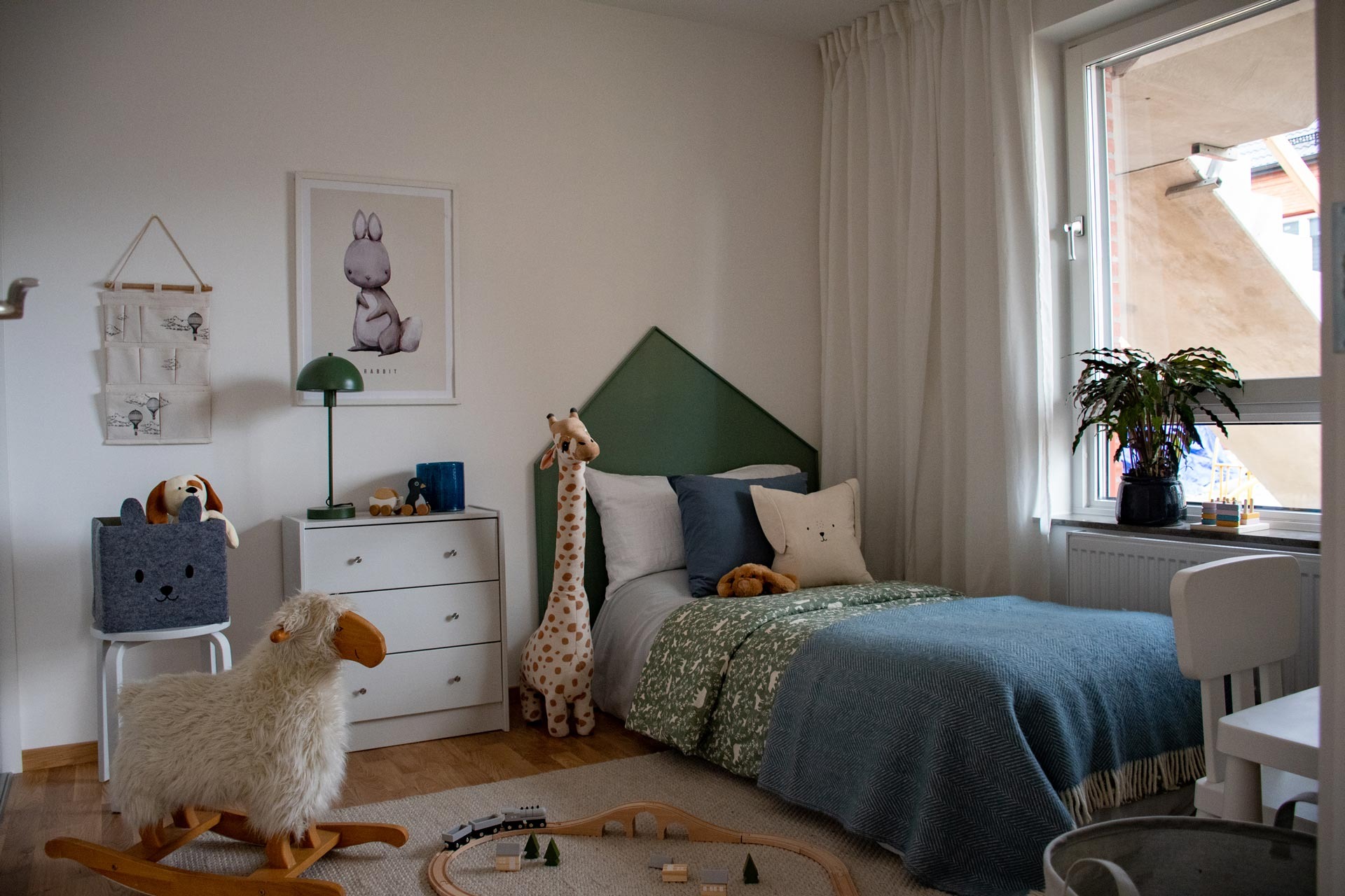 Ett barnrum som är väl upplyst och mysigt, med ett stort fönster till höger. Rummet har en enkelsäng med blågrönt överkast och flera mjukdjur på den. Till vänster om sängen finns en vit byrå med en leksaksgiraff bredvid, och ovanför byrån hänger en illustration av en djurfigur. På rummets vänstra sida finns ett barnanpassat bord och stolset, med en gungande fårleksak framför.