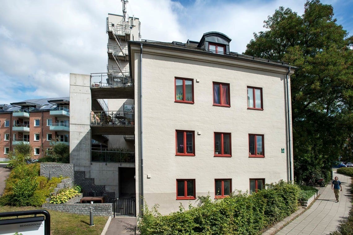 En vit byggnad med röda fönsterkarmar, omgiven av grönska och en gångväg. Bakgrunden pryds av träd och en blå himmel med vita moln.