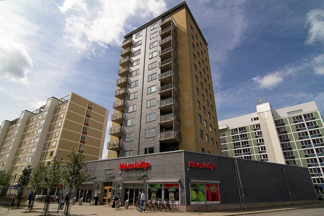 En modern stadsdel med flera höghus och en butik vid namn “Hemköp” på gatunivå. Det är en klar dag, och byggnaderna har en samtida arkitektonisk design.