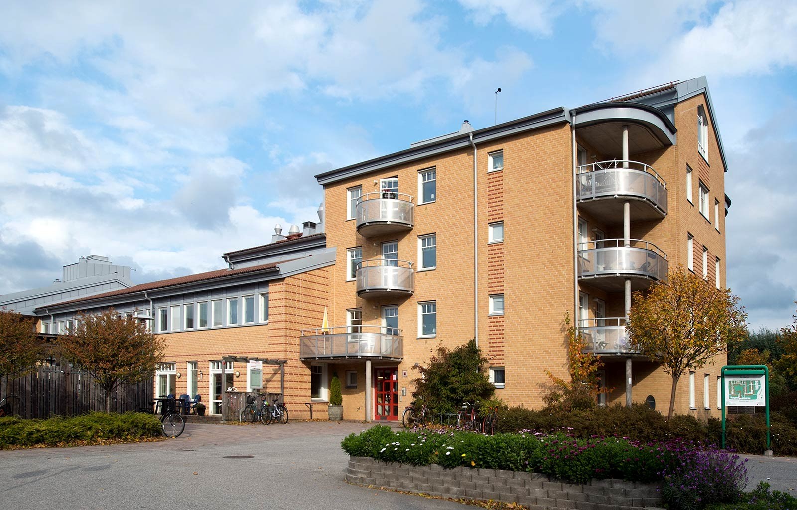 En gul tegelbyggnad med tre våningar och rundade balkonger på höger sida. Det finns cyklar parkerade framför byggnaden och en liten trädgård med blommor och buskar är synlig.