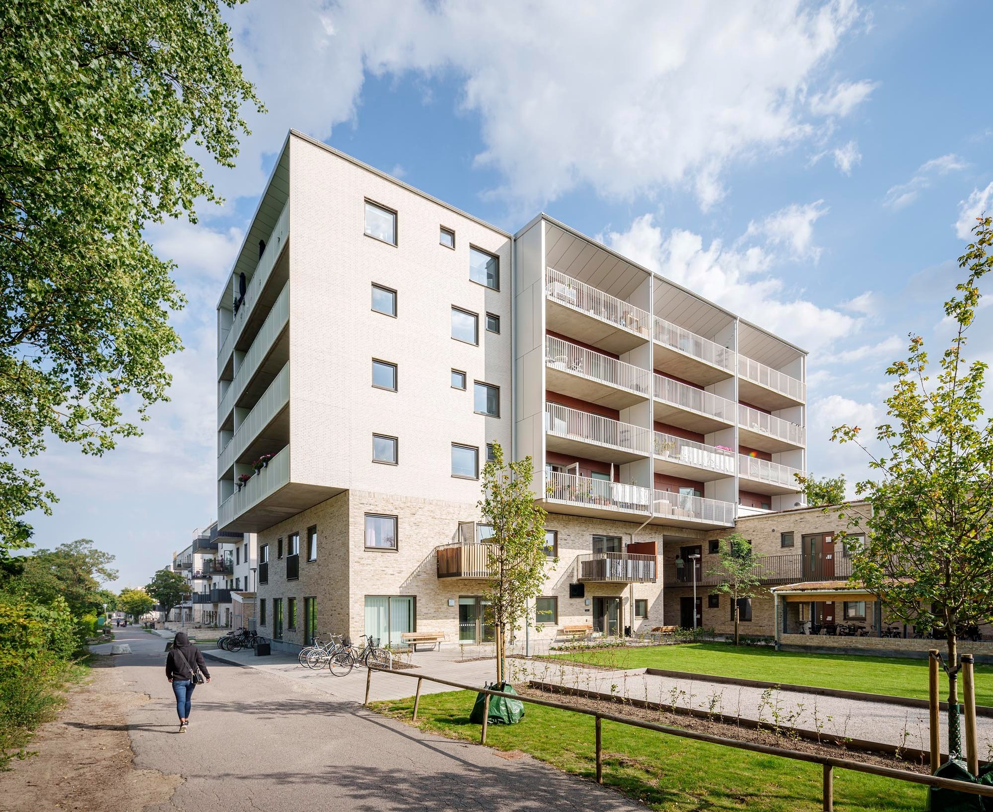 En modern flervåningsbyggnad med vit fasad och flera balkonger. Grönska inklusive gräs och träd, samt en klar himmel.