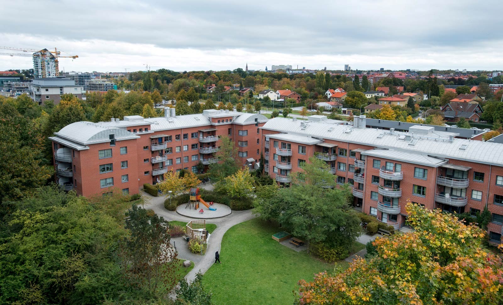 En utsikt över ett bostadsområde med röda tegelbyggnader, en lekplats, och träd med höstfärger. En större stadssilhuett syns i bakgrunden. Atmosfären är lugn och familjevänlig.