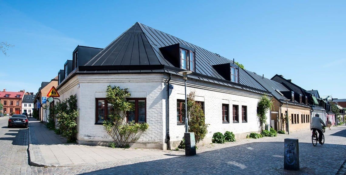 En lugn gata med en vit byggnad, vars tak är prytt av svarta takplattor. En person på cykel och en bil syns i bilden.