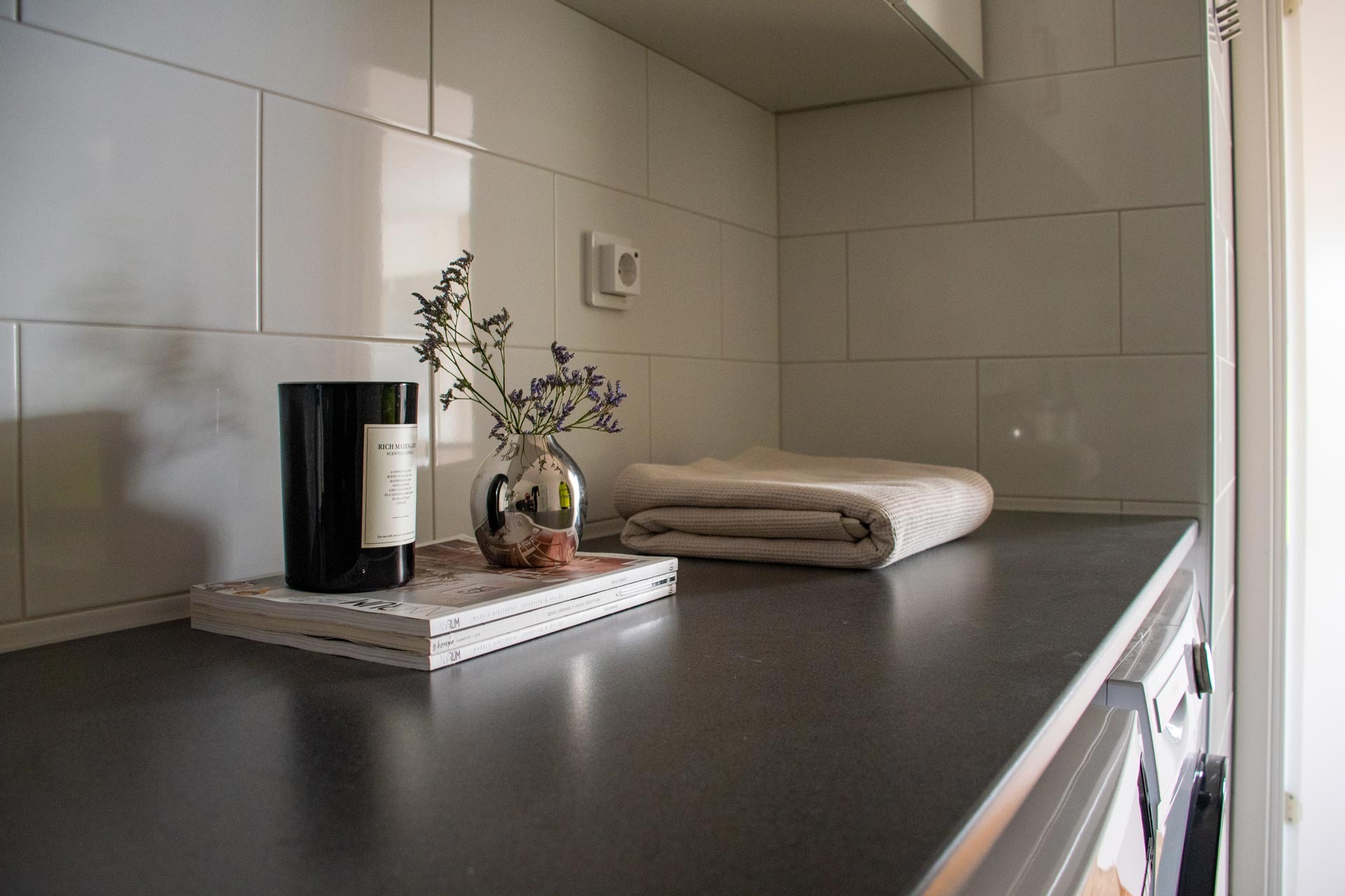 En snyggt organiserad köksbänk med en mörk yta. På bänken finns en hög med vita böcker eller tidskrifter som är toppade med en svart cylindrisk behållare, bredvid vilken det står en liten glasvas med lila blommor. Bredvid dessa föremål finns en snyggt vikt ljusfärgad kökshandduk.